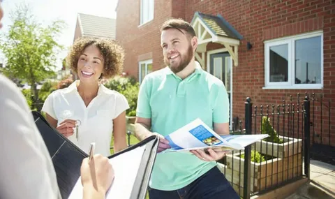 A smiling couple stands in front of a brick house while a person in the foreground writes in a folder, holding keys and a brochure during a meeting about mortgage options for buying a home.